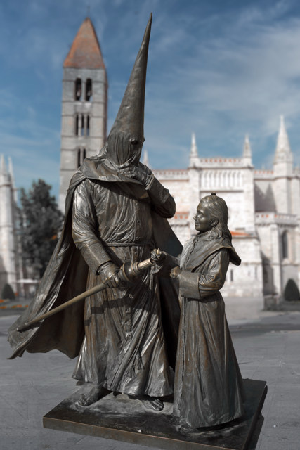 Escultura de un penitente con capirote junto a una figura infantil que alza las manos, con una iglesia desenfocada detrás en Valladolid.
