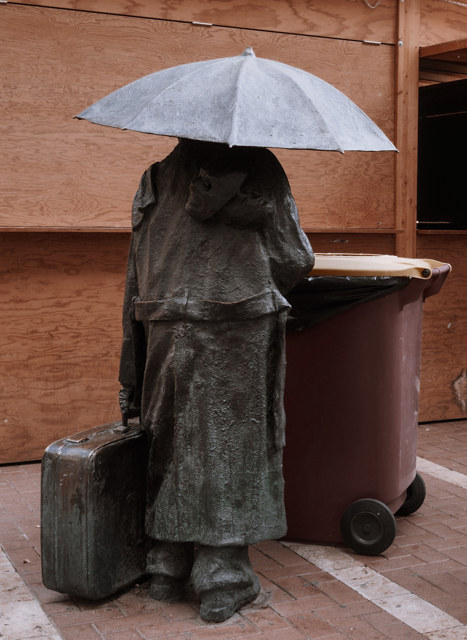 Estatua sentada bajo una sombrilla junto a un cubo de basura, ante un cerramiento de madera en Valladolid.