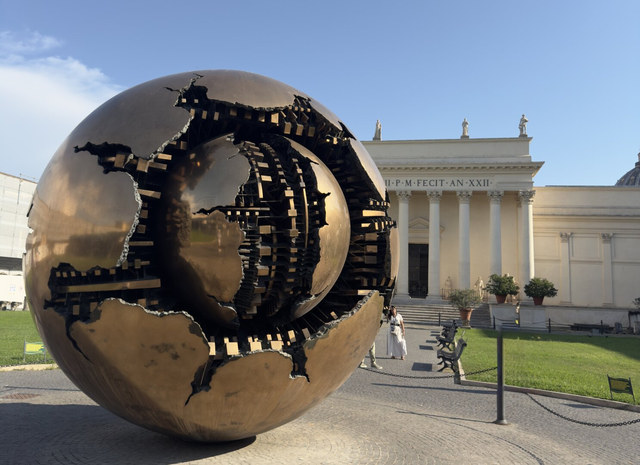 Una esfera de bronce agrietada revela otra esfera interior frente a una fachada neoclásica del Vaticano.