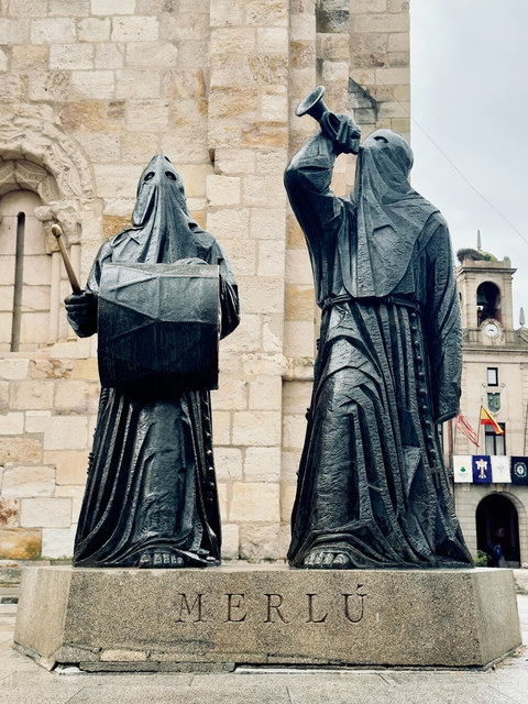 Dos figuras encapuchadas de bronce sostienen un tambor y una corneta frente a una iglesia de Zamora.
