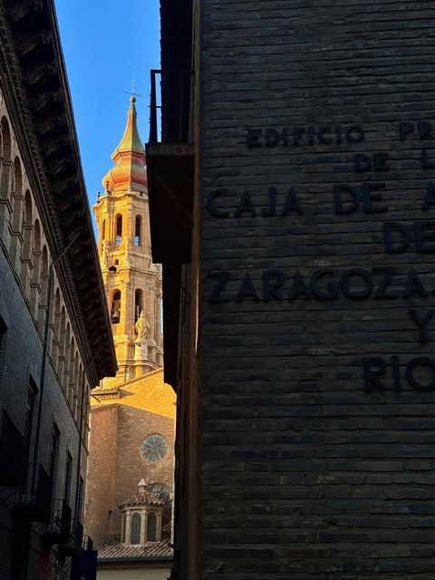 Una torre barroca iluminada aparece entre dos fachadas oscuras de ladrillo en una calle estrecha de Zaragoza.